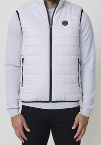 Homme portant un gilet matelassé gris clair à fermeture éclair par-dessus une chemise texturée gris clair à manches longues et un pantalon noir, debout avec les mains détendues.