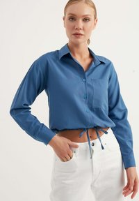 Camicia blu a maniche lunghe con colletto, chiusura frontale con bottoni e taschino sul petto. Jeans bianchi con vita legata, texture liscia.