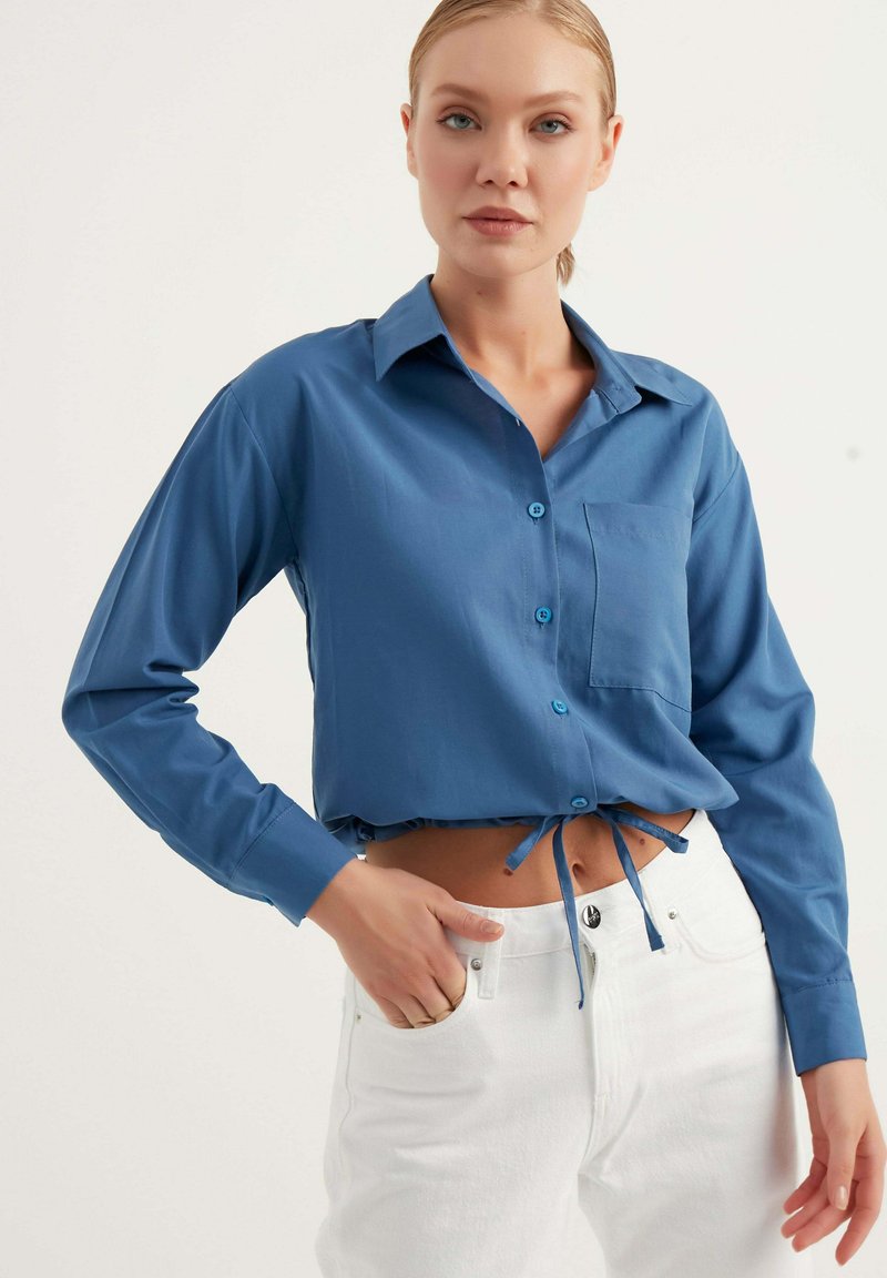 Camicia blu a maniche lunghe con colletto, chiusura frontale con bottoni e taschino sul petto. Jeans bianchi con vita legata, texture liscia.