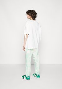 Camiseta blanca de gran tamaño con textura suave y lazos en los laterales, combinada con pantalones de chándal verde menta de tie-dye. Zapatillas verdes y blancas completan el look.