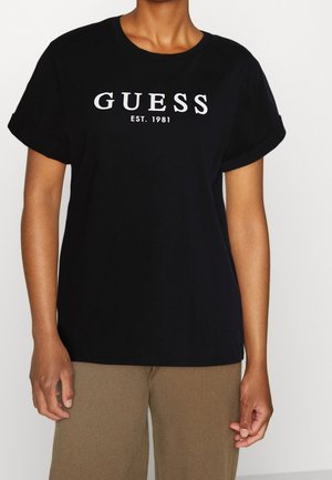 Personne portant un t-shirt noir à manches courtes avec le texte "GUESS EST. 1981" en blanc, assorti à un pantalon marron clair.
