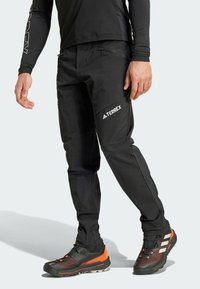 Černé outdoorové kalhoty s úzkým střihem, zipovými kapsami a texturovaným povrchem, v kombinaci s vícebarevnými sportovními botami.