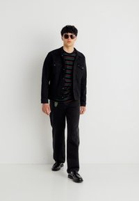 Νεαρός άνδρας που φορά μαύρο τζιν μπουφάν, ριγέ πουκάμισο, μαύρο παντελόνι, μαύρα παπούτσια και γυαλιά ηλίου, στέκεται μπροστά σε λευκό φόντο.