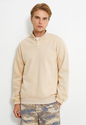 Ljust beige hoodie med upphöjd krage, tryckknappar och ribbade mudd. Bärs med mönstrade shorts i nyanser av blått och beige.