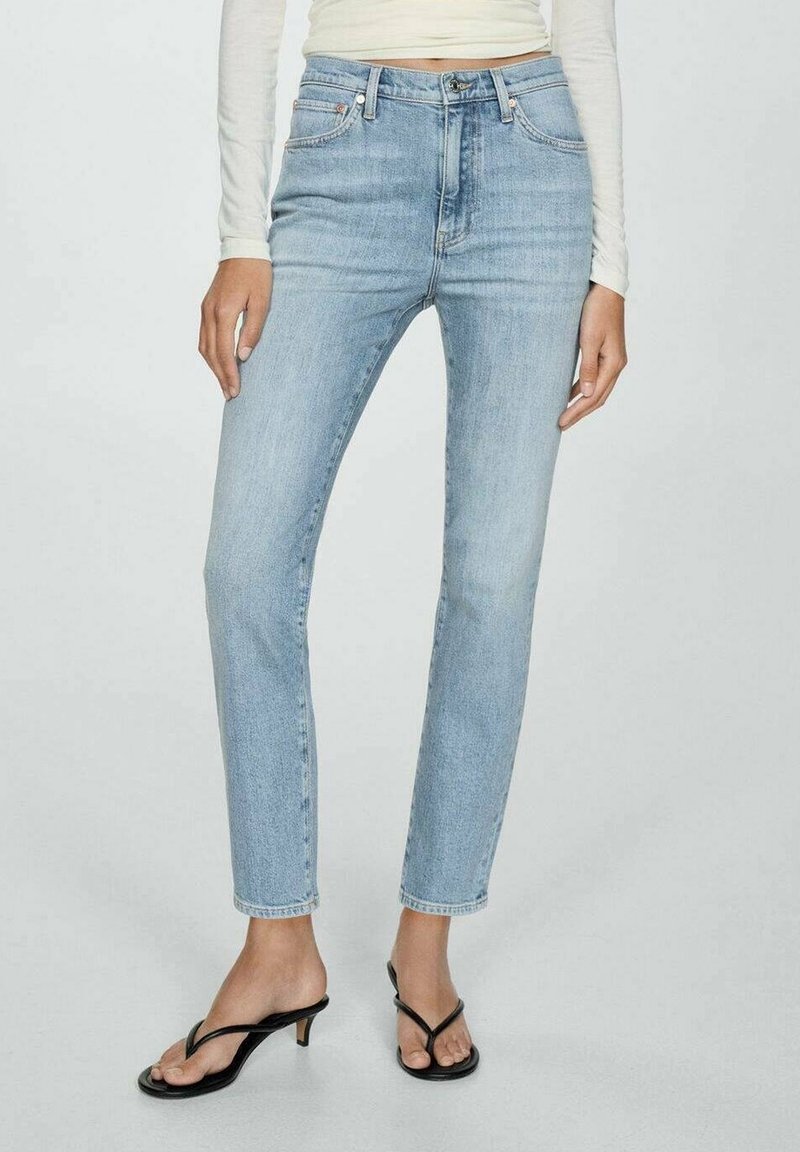 Personne portant un jean slim taille haute bleu clair, un top court blanc à manches longues et des sandales noires à talons, debout devant un fond uni.