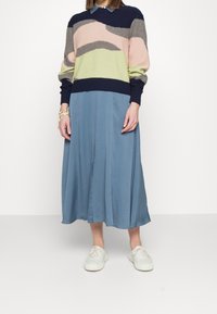 Personne portant un pull à motifs multicolores, un pantalon large bleu, des baskets blanches et des bracelets colorés en perles au poignet gauche.