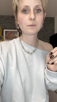 Svijetlosivi oversized dukserica s dizajnom koji otkriva jedno rame. Debela srebrna ogrlica s mašnama. Kratki crni nokti s gradijentnim dizajnom.