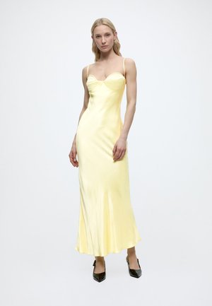 Ženska stoji in nosi dolgo bledo rumeno satensko obleko z tankimi naramnicami ter črne koničaste pete na belem ozadju.