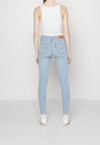 Jeans skinny de color azul claro con cintura alta, costuras en los bolsillos traseros con un diseño curvado, combinados con una camiseta sin mangas blanca y zapatos deportivos.