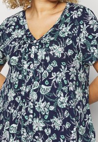 Top de patrón florido en color azul marino con acentos en verde y blanco; presenta un escote en V, mangas cortas y detalles de botones en el frente.