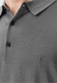 Nærbillede af en person iført en grå strikket polo-shirt med krave, to sorte knapper og et lille broderet gedeskull logo på brystet.