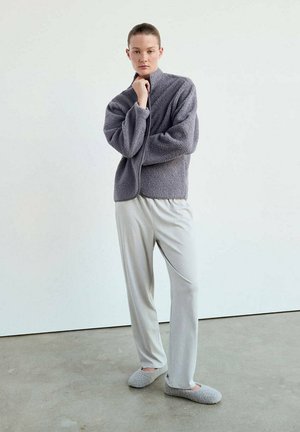 Persona joven está de pie vistiendo una chaqueta de forro polar gris, pantalones sueltos gris claro y zapatillas grises a juego sobre un suelo de hormigón frente a una pared blanca.
