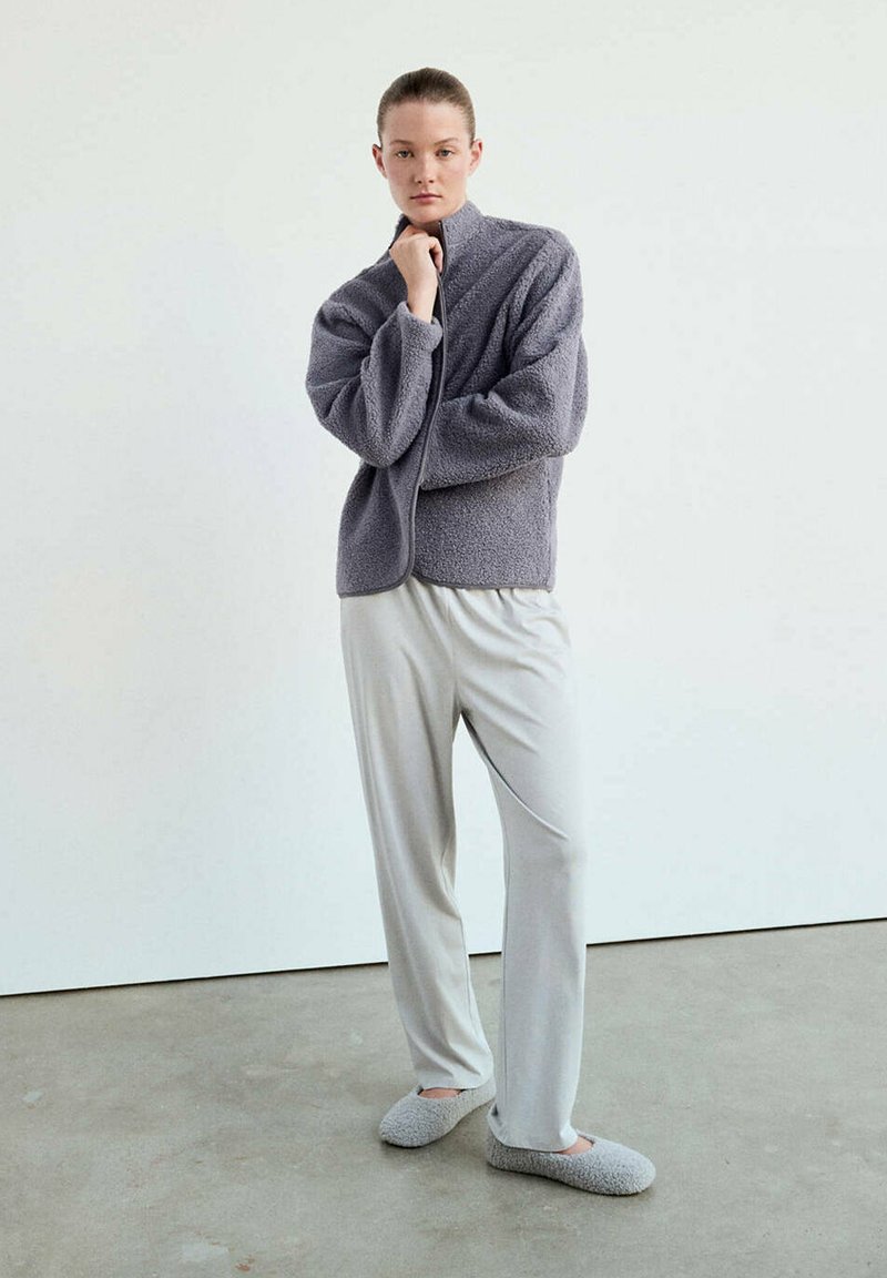 Jeune personne debout portant une veste polaire grise, un pantalon ample gris clair et des chaussons gris assortis sur un sol en béton contre un mur blanc.