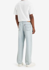 Uomo in piedi con indosso jeans diritti azzurri chiari, una maglietta corta bianca e sneakers bianche, rivolto di spalle su uno sfondo bianco.