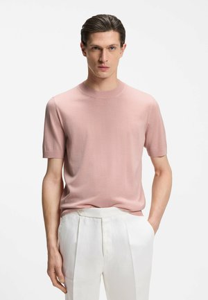 Mężczyzna z zaczesanymi do tyłu ciemnymi włosami, ubrany w jasnoróżowy sweter z krótkim rękawem i białe spodnie z wysokim stanem, z rękami w kieszeniach.