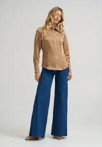 Jasno brązowa satynowa koszula na guziki z wywijanym kołnierzykiem. Połączona z rozszerzającymi się ciemnoniebieskimi dżinsami oraz metalicznymi złotymi butami.