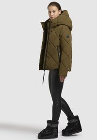 Olijfgroene gewatteerde pufferjas met een hoge kraag, rits aan de voorkant en zwarte accenten, gecombineerd met zwarte neplederen leggings en laarzen.