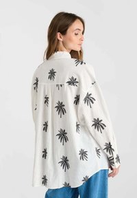 White linen shirt featuring black palm tree patterns, long sleeves, relaxed fit, and a high collar, paired with blue jeans.