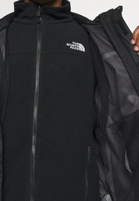Czarna kurtka z miękkiego materiału polarowego, z zamkiem z przodu, wysokim kołnierzem, bocznymi kieszeniami, siateczkową podszewką i białym logo „The North Face”.
