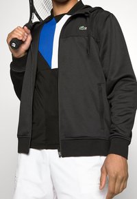 Svart blixtlåströja med huva, med en grön logotyp. Under den en svart och blå sporttröja. Vita shorts och en tennisracket i handen.