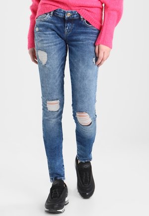 Personne portant un jean skinny bleu déchiré, des baskets noires et un pull rose vif, debout devant un fond clair uni.