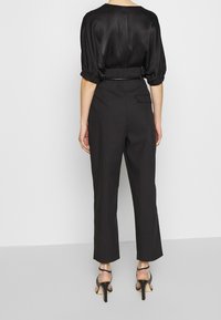 Blusa nera di satin con maniche a sbuffo, abbinata a pantaloni neri a vita alta con tasca posteriore e taglio slim alla caviglia. Tacchi a strappo neri.