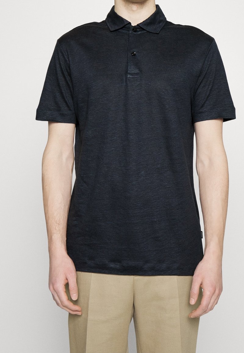Homme portant un polo manches courtes bleu marine foncé à deux boutons et un pantalon beige, sur un fond uni.