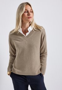 Beige v-neck sweater made of soft knit fabric, featuring ribbed cuffs and hem, worn over a white collared shirt, with a relaxed fit.