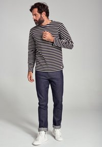 Langærmet skjorte i marineblå og cremefarvede striber, parret med mørke jeans og hvide sneakers, som viser en afslappet pasform med opkrøllede manchetter.