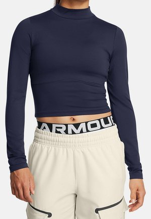 Vrouw draagt een navy lange mouwen cropped top en beige broek met een zichtbare zwart-witte "ARMOUR" tailleband.