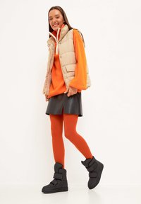 Vesta puf bej, pulover supradimensionat portocaliu, fustă din piele neagră, ciorapi portocalii și botine negre cu cataramă. Texturi netede și design stratificat.