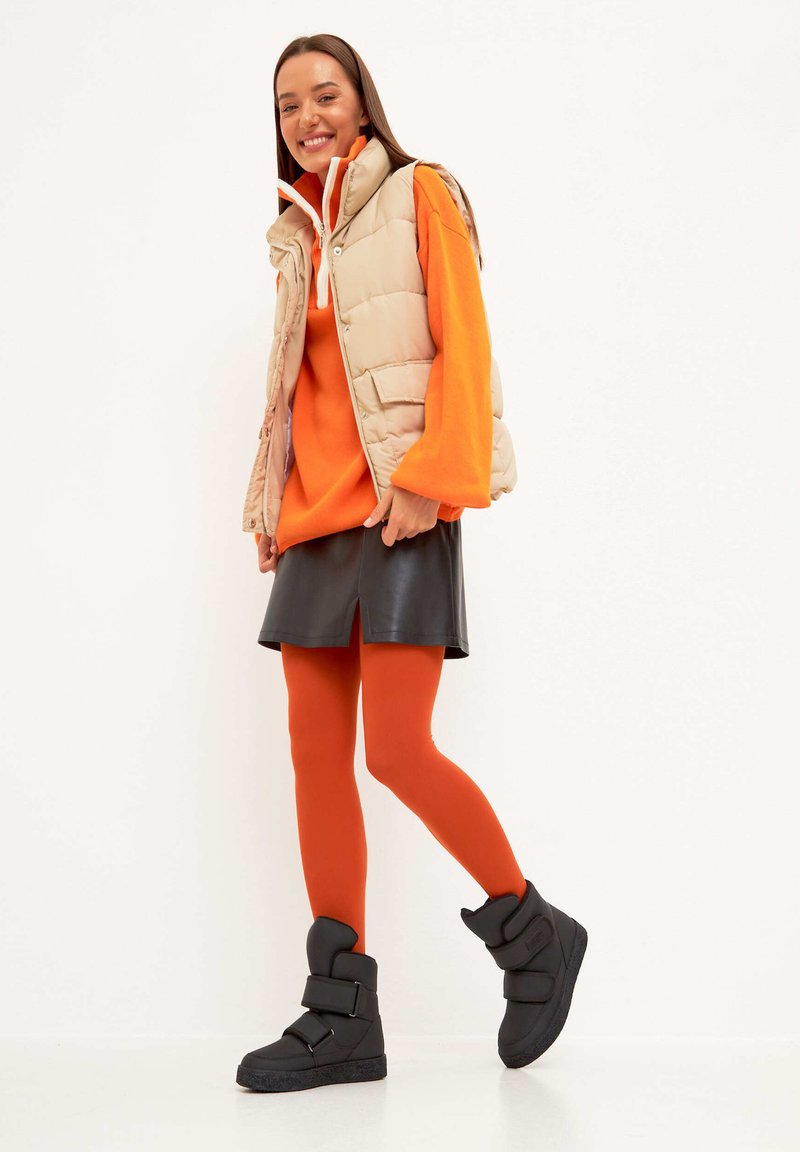 Vesta puf bej, pulover supradimensionat portocaliu, fustă din piele neagră, ciorapi portocalii și botine negre cu cataramă. Texturi netede și design stratificat.