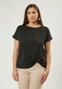 Top in satin nero con maniche corte, scollo rotondo e dettaglio a nodo sul lato inferiore, abbinato a pantaloni beige. Texture semplice e liscia.