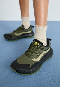 Baskets Vans vertes et noires avec une tige texturée, arborant un design à rayures beige, des lacets ronds et une semelle en caoutchouc avec des accents verts.