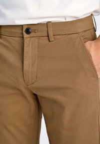 Bruna bomullsblandade byxor med rak passform, dragkedja och knappstängning, sidofickor och en slät yta. Synlig sömsdetalj.