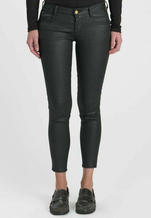 Donna che indossa pantaloni aderenti in pelle nera e mocassini slip-on neri, in piedi contro uno sfondo chiaro semplice.
