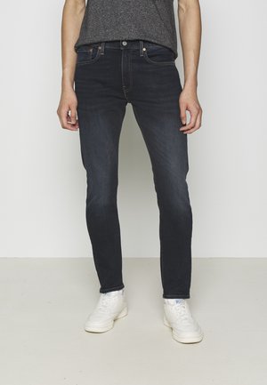 Jeans ajustados de color azul oscuro fabricados en denim, con cierre estándar de botón y cremallera. Combinados con zapatillas blancas y una camiseta gris.