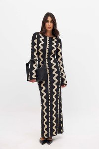 Robe noire à manches longues avec un motif ondulé crème, silhouette cintrée, tissu en tricot texturé. Le modèle tient un sac en cuir noir. Chaussures noires à talons pointus.