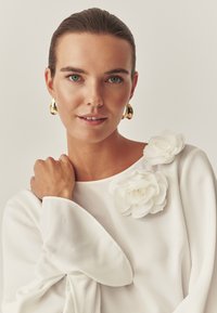 Biała bluzka z miękkiego materiału, zdobiona dwoma kwiatowymi akcentami na ramieniu oraz szerokimi, zwiewnymi rękawami. Złote kolczyki w kształcie kół.