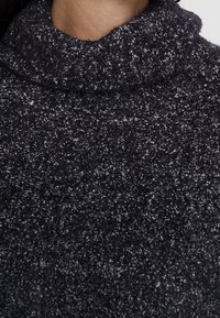 Marineblauwe gebreide trui met een hoge, opgerolde kraag. De textuur is pluizig en zacht, met een gespikkeld patroon in lichtere tinten.