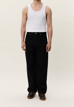 Homme portant un débardeur blanc sans manches, un pantalon noir ample, et des chaussures marron foncé, debout devant un fond clair uni.