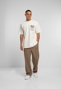 Krémové oversized tričko s krátkým rukávem a grafickým potiskem na přední straně, spárované s hnědými kalhotami a bílými teniskami.
