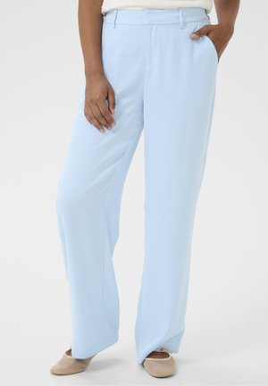 Persona que lleva pantalones rectos azul claro con trabillas para cinturón, camiseta blanca, zapatos beige sin cordones, una mano en el bolsillo y la otra descansando sobre el muslo.