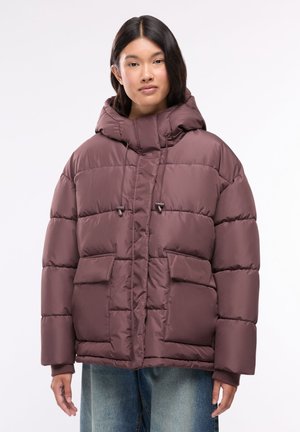 Donna con giacca imbottita viola con cappuccio, grandi tasche anteriori e jeans blu, in piedi davanti a uno sfondo bianco uniforme.