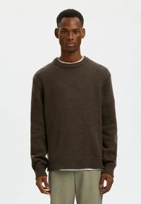 Maglione marrone a maglia con scollo rotondo, polsini e orlo a coste. Presenta una vestibilità rilassata e una superficie testurizzata. Indossato sopra una camicia bianca.