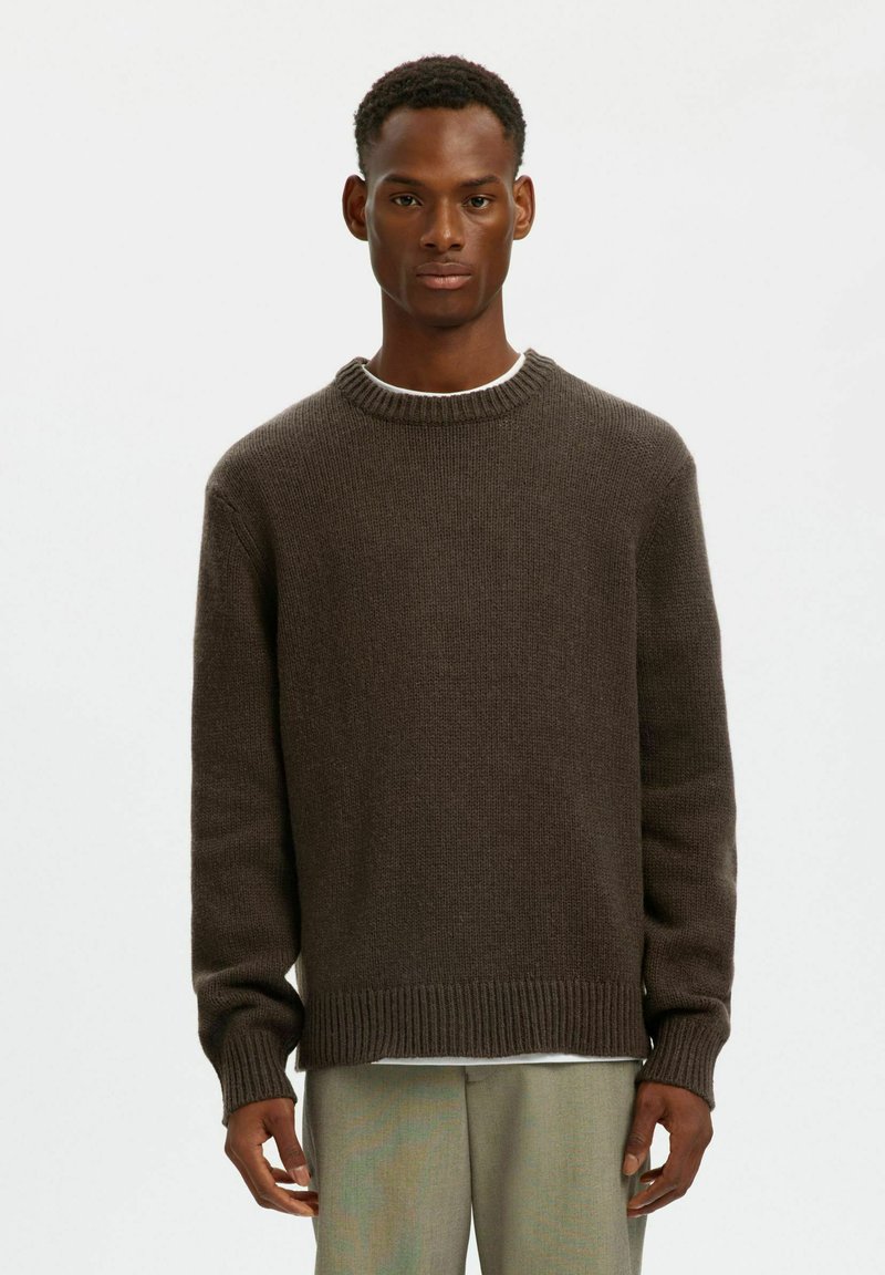 Maglione marrone a maglia con scollo rotondo, polsini e orlo a coste. Presenta una vestibilità rilassata e una superficie testurizzata. Indossato sopra una camicia bianca.