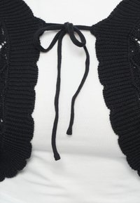 Μαύρο πλεγμένο στρογγυλό πλεκτό με κυματοειδείς άκρες που δένεται με φιόγκο πάνω από λευκό ριμπ πουκάμισο.