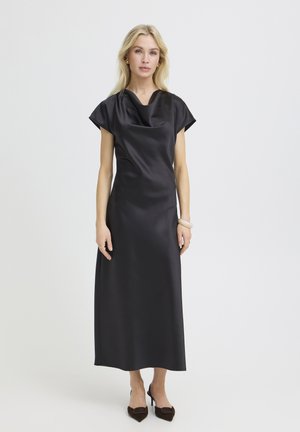 Blondínka v čiernych saténových midi šatách bez rukávov s výstrihom na krku a čiernymi lodičkami s ostrou špičkou stojaca pred bielym pozadím.