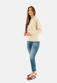 Felpa con zip beige dal tessuto morbido, con cappuccio e maniche raglan. Abbinata a jeans slim blu chiaro e scarpe sportive color bordeaux.