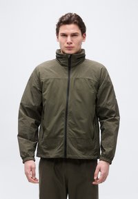 Νεαρός άνδρας που φοράει μπουφάν με κουκούλα σε ελαιοπράσινο χρώμα, με φερμουάρ μπροστά και υφή στα μανίκια και τους ώμους, στέκεται μπροστά σε απλό φόντο.